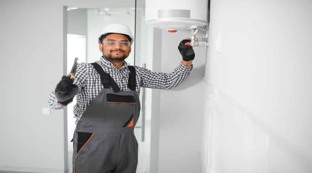 Indian plumber using screwdriver while fixing boiler or water heater, working on heating system in apartment