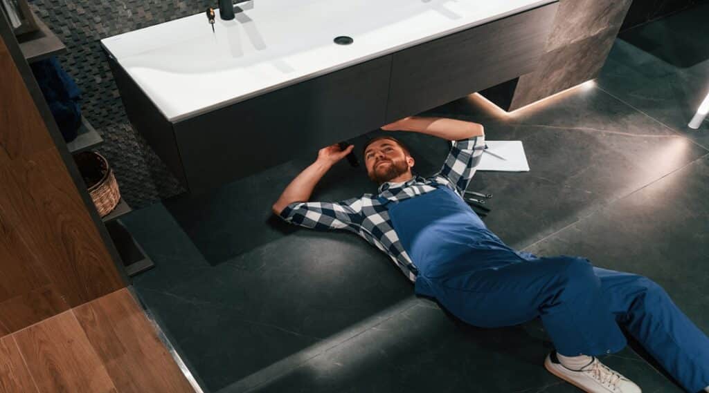 Fixing the siphone. Lying down on the floor. Plumber in blue uniform is at work in the bathroom